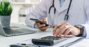 Doctor in a white coat using a calculator while holding a smartphone, with a laptop and medical documents on the desk.