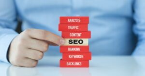 Hand pointing to a wooden block labeled SEO in a stacked tower of red blocks with words analysis, traffic, content, ranking, keywords, and backlinks.