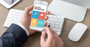 A person in a business suit holding a smartphone displaying a “Digital Marketing” screen with colorful sections labeled SEO, Content, Research, and Viral, while sitting at a desk with a keyboard, computer monitor, and wireless mouse.