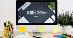 A desktop computer on a wooden desk displaying a digital agency website with text about web design, digital marketing, and creative design. The desk is decorated with colorful stationery, sticky notes, notebooks, and potted plants.