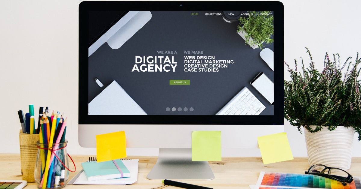 A desktop computer on a wooden desk displaying a digital agency website with text about web design, digital marketing, and creative design. The desk is decorated with colorful stationery, sticky notes, notebooks, and potted plants.