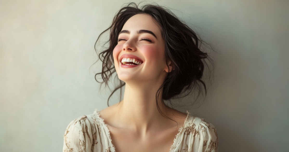 A young woman with dark, loosely styled hair smiles broadly with her eyes closed, head tilted slightly back, wearing a light, delicate blouse against a soft, neutral background.