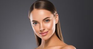 Studio portrait of a young woman with smooth, glowing skin, blue eyes, and straight brown hair parted in the center, wearing natural makeup and a subtle smile against a dark gradient background.