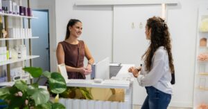 A medspa receptionist smiles while assisting a client at the front desk, with skincare products displayed on shelves and a clean, modern clinic interior in the background.