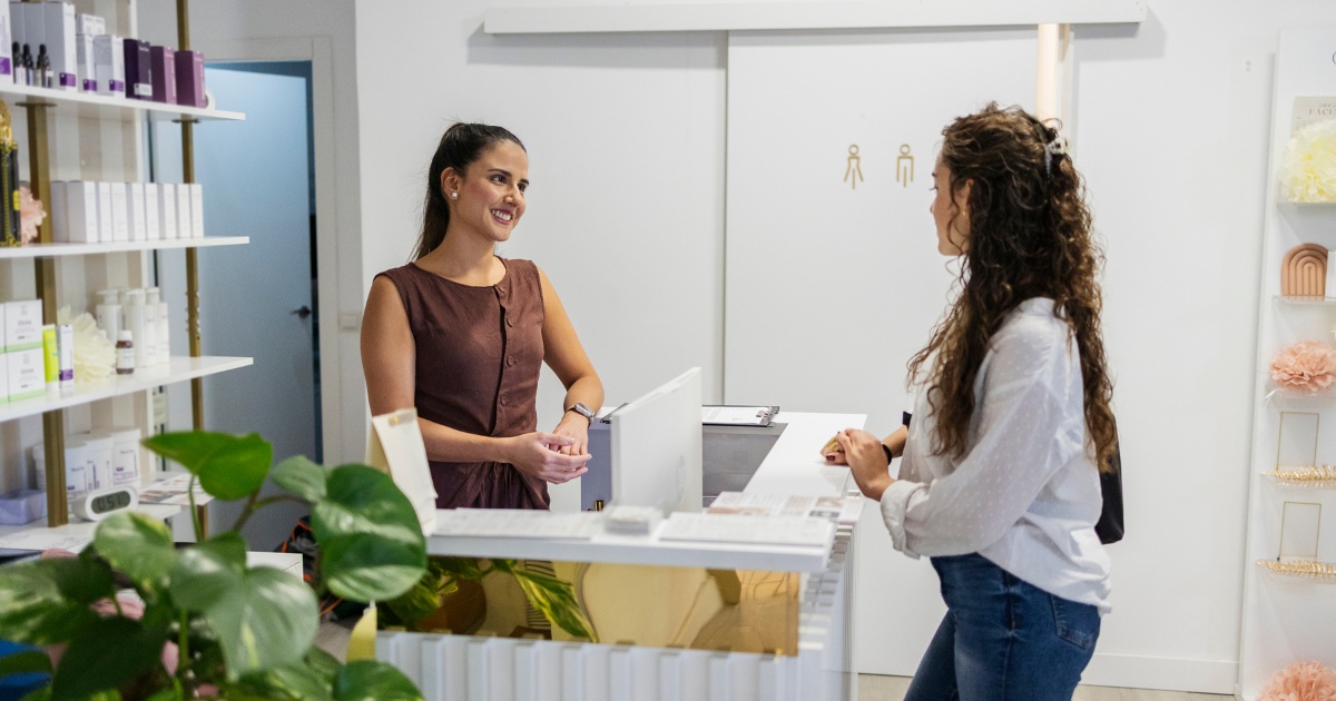 A medspa receptionist smiles while assisting a client at the front desk, with skincare products displayed on shelves and a clean, modern clinic interior in the background.