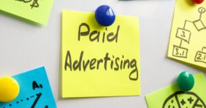 Yellow sticky note with the handwritten words “Paid Advertising” pinned to a board with a blue pushpin, surrounded by colorful notes showing charts and marketing diagrams.