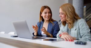 Two women discussing work while looking at a laptop and tablet in an office setting.