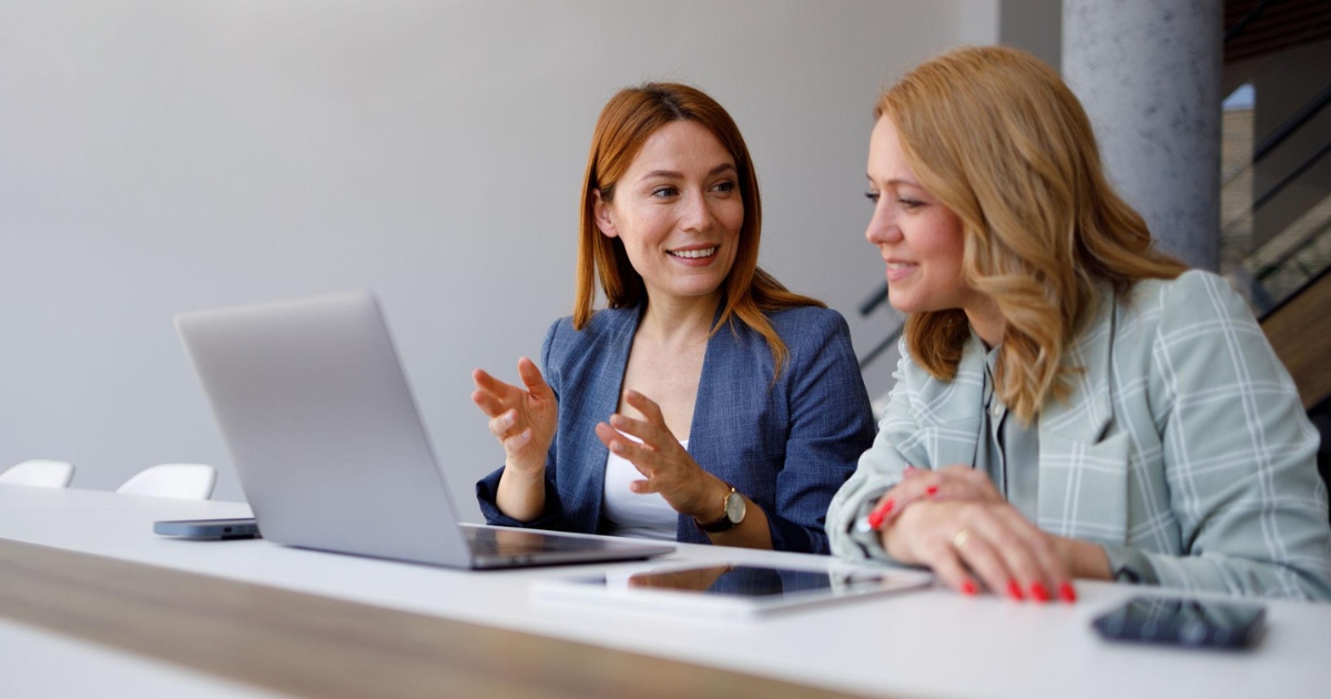 Two women discussing work while looking at a laptop and tablet in an office setting.